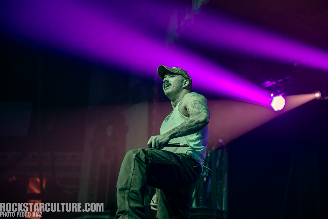 A musician performing on stage under colorful lighting, wearing a tank top and a cap, passionately singing into a microphone.