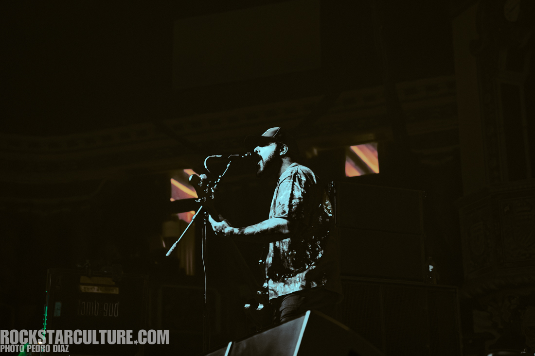 A musician passionately performing on stage with a guitar, singing into a microphone, surrounded by dim lighting and a concert atmosphere.