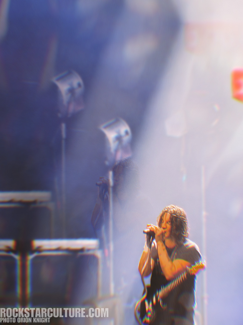 A musician performing on stage, holding a microphone and guitar, with bright stage lights in the background.