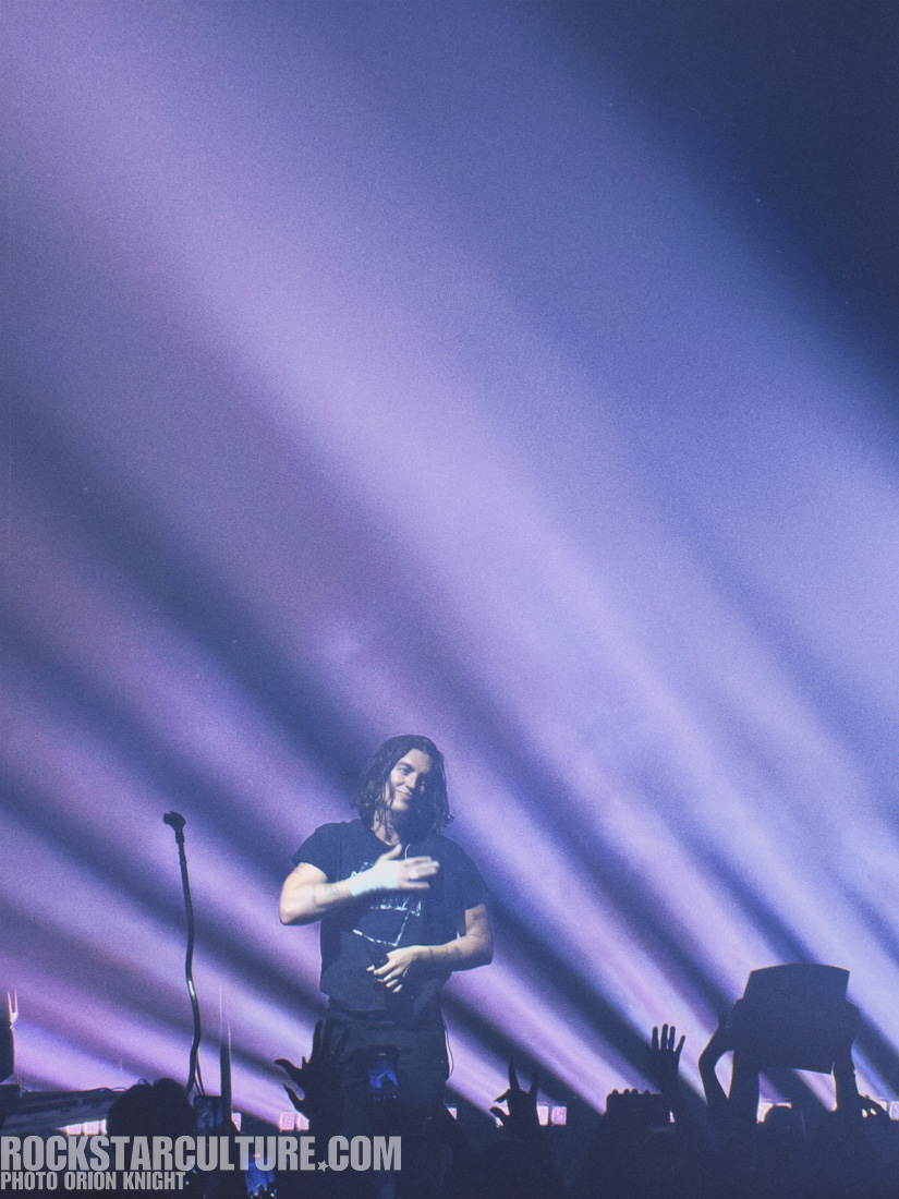 A performer on stage with long hair, wearing a black t-shirt, interacting with the audience under vibrant purple stage lighting.