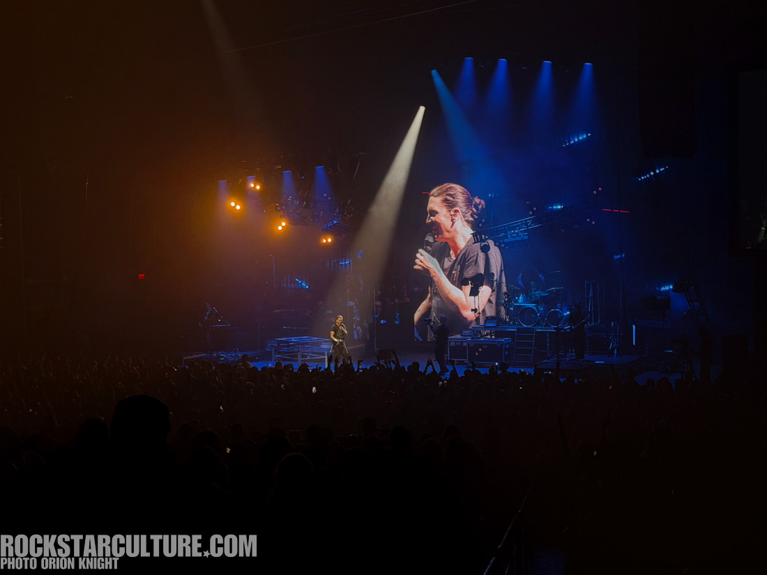 A singer performing on stage with a large screen behind displaying their image, surrounded by colorful stage lights and a cheering crowd.