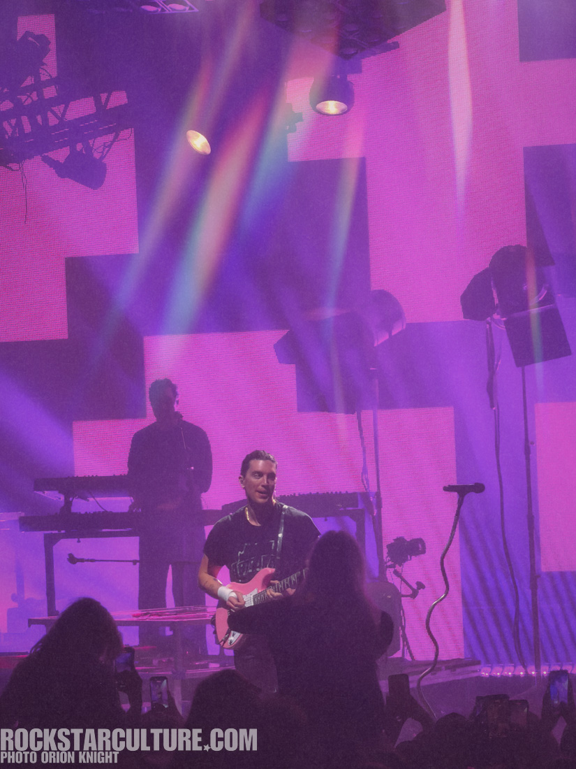 A guitarist performing on stage with a pink guitar, illuminated by colorful stage lights against a purple background, while an audience member captures the moment on their phone.