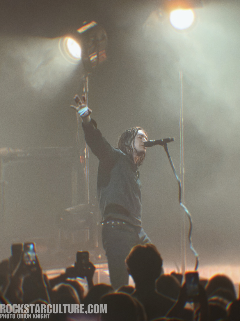 A performer with long hair stands on stage, passionately singing into a microphone while raising one hand. Bright stage lights illuminate the scene, surrounded by an audience holding up their phones.