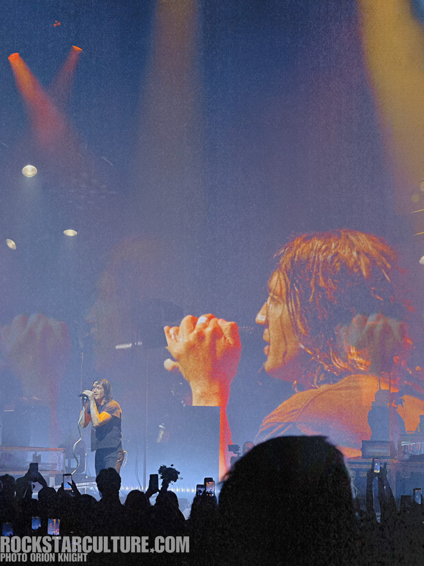 A singer performing on stage with dramatic lighting and a large screen displaying their image in the background, viewed by an audience holding smartphones.