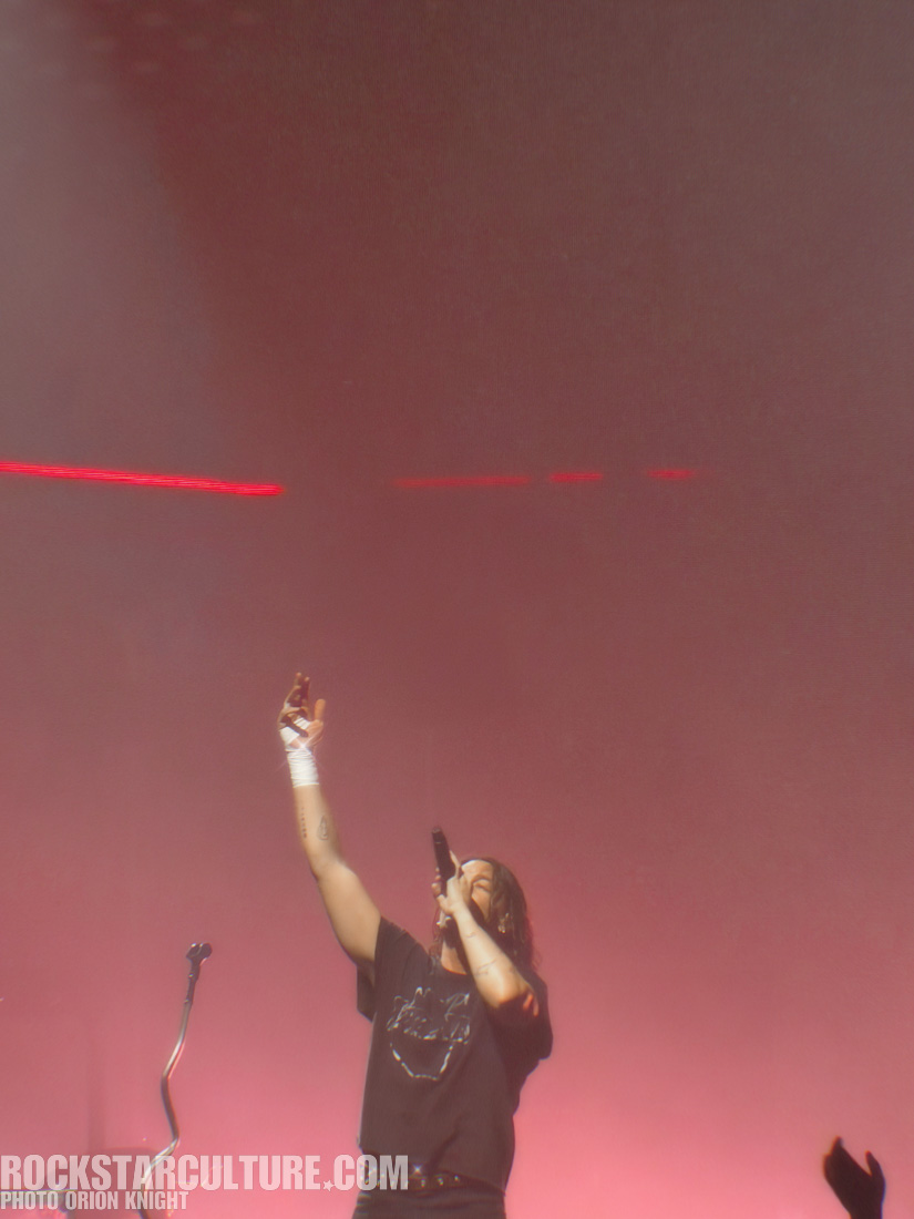 A singer performing on stage with one arm raised, wearing a black t-shirt and wrapped bandages on their hands, with a colorful light backdrop.