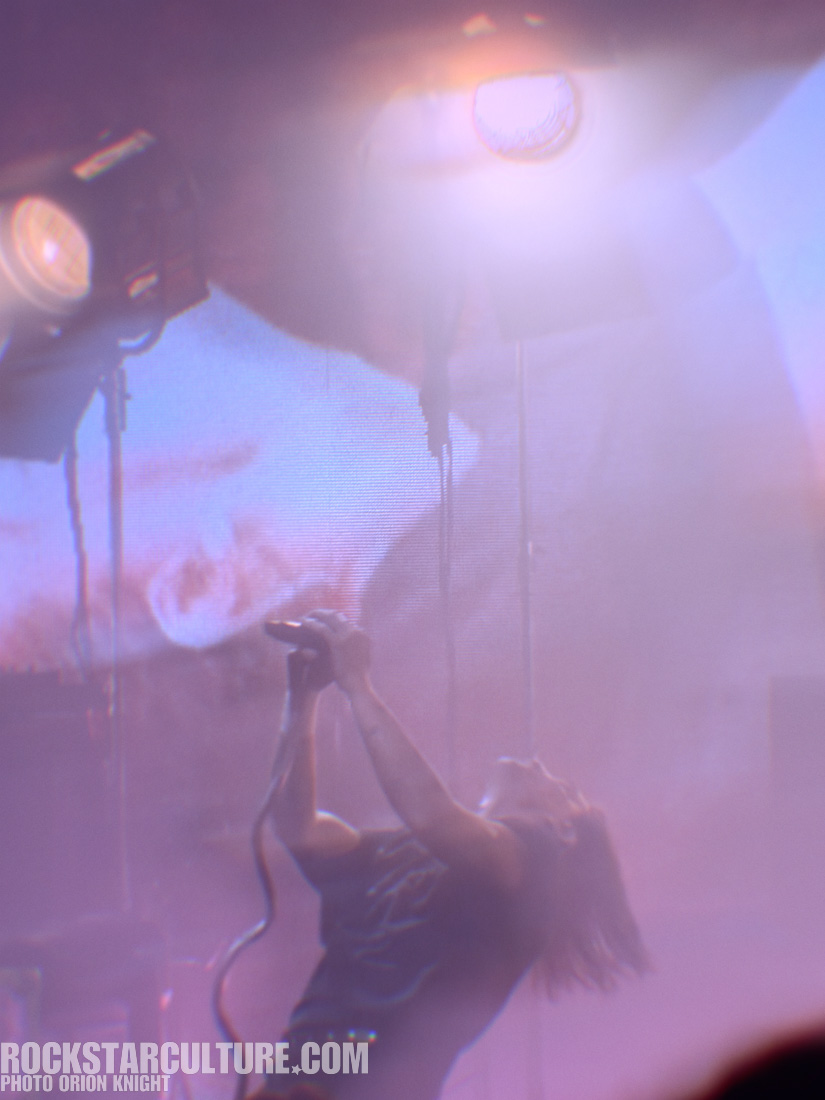 A performer with long hair passionately singing into a microphone on stage, illuminated by bright stage lights, with a blurred background displaying visuals.