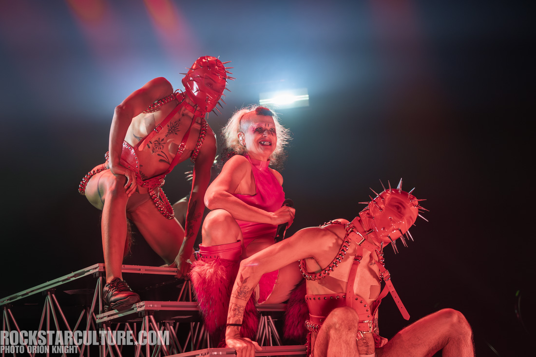 Three performers on stage, with one wearing a bright pink outfit and two others in leather harnesses with spikes, posing dramatically under stage lights.
