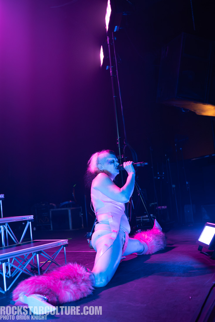 A performer kneeling on stage while singing into a microphone, wearing a colorful outfit with furry accents, illuminated by purple stage lights.