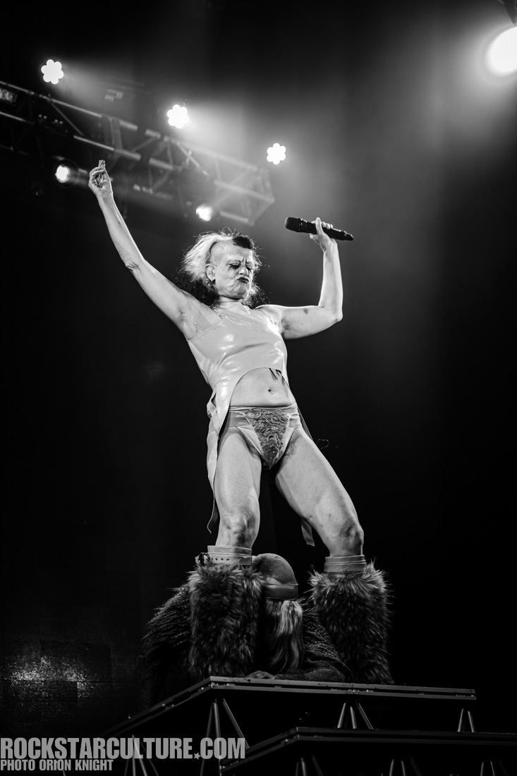 A performer on stage striking a dramatic pose while holding a microphone. The performer is dressed in a unique outfit with a shiny top and revealing bottoms. The background features stage lighting and a sense of energy and excitement.