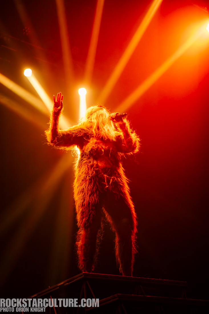 A performer dressed in a furry costume stands on a raised platform, singing into a microphone under dramatic orange spotlighting at a concert.