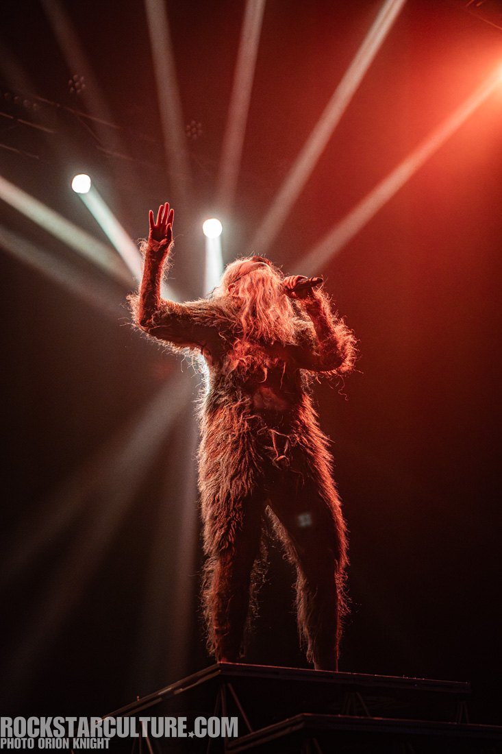 A performer wearing a furry costume and long hair sings passionately on stage, with dramatic lighting creating a dynamic atmosphere.