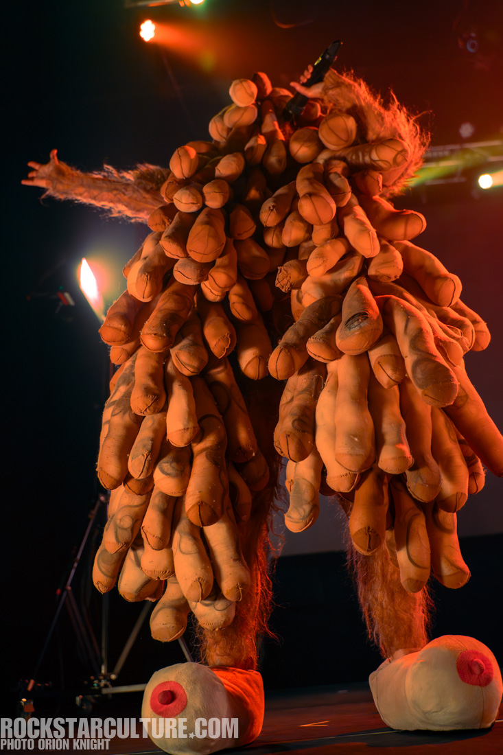 A performer wearing a costume covered with multiple plush limbs, holding a microphone, in a stage setting with colorful lighting.