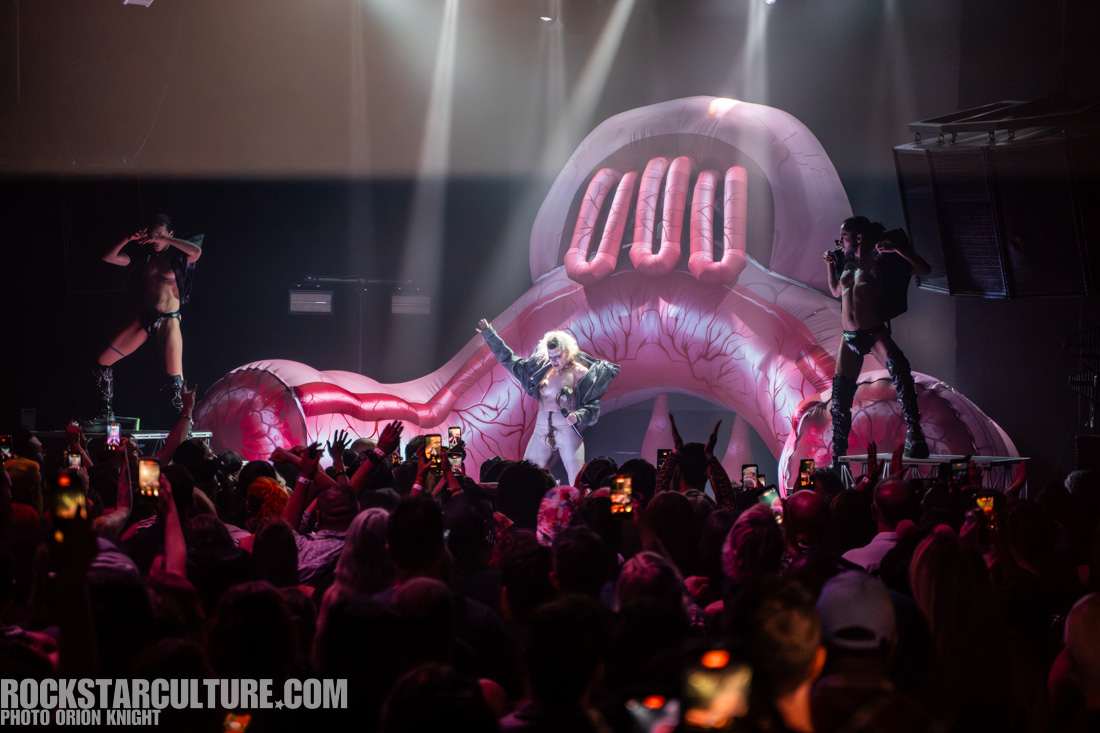 A performer in a shiny outfit stands on stage, singing into a microphone with a large inflatable pink set of lips and teeth in the background. Two dancers pose on either side of the performer, while an engaged audience captures the moment with their phones.