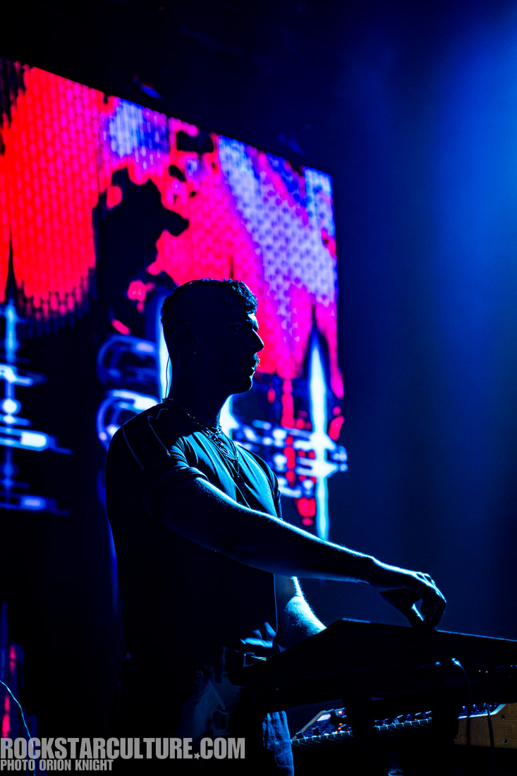 A musician performing on stage, silhouetted against a vibrant, colorful light display, playing a synthesizer or keyboard.