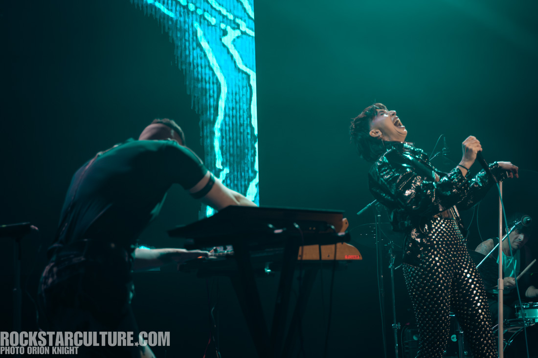 A performer singing passionately on stage while another musician plays a keyboard in the background, with colorful lighting and digital visuals creating an energetic atmosphere.