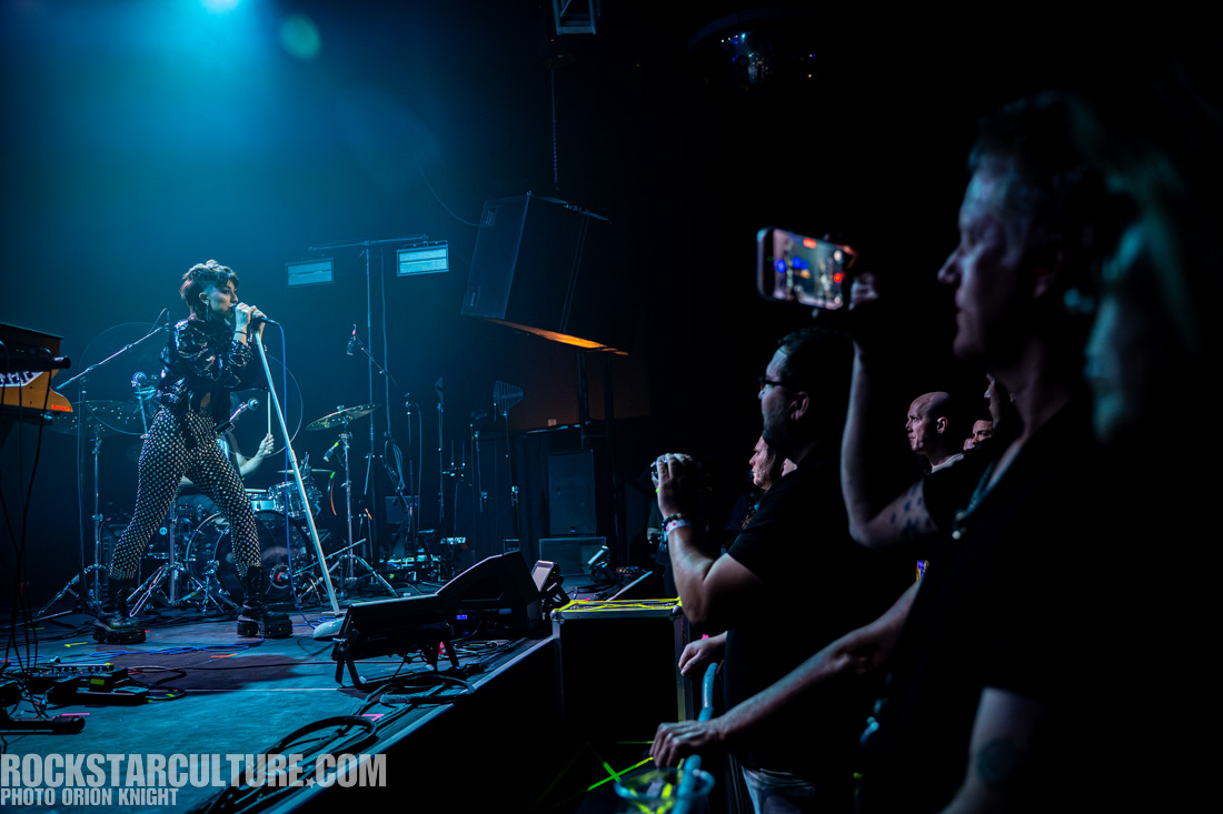 A performer singing into a microphone on stage, dressed in a shiny black outfit with patterned pants, while the audience watches and some capture the moment on their smartphones.