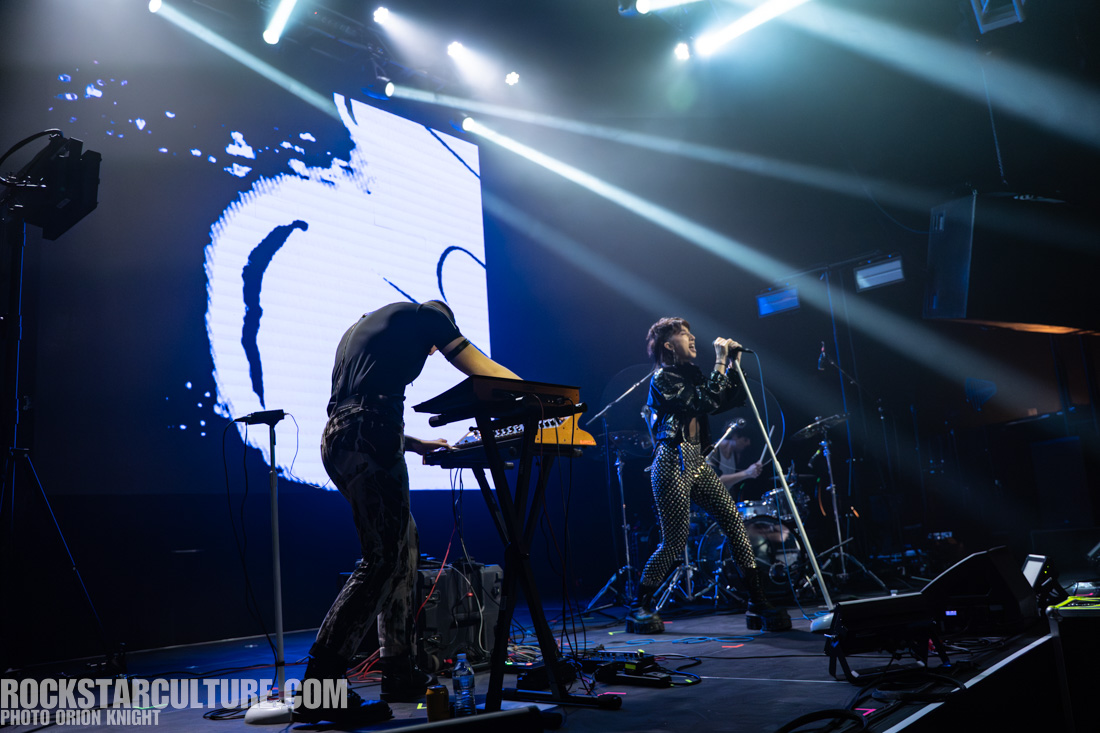 A live music performance with a singer using a microphone and a musician playing a keyboard, illuminated by dynamic stage lights and a visual backdrop.