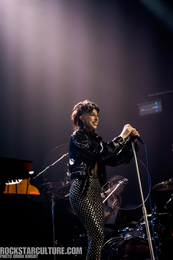 A performer on stage smiling and holding a microphone, wearing a shiny black jacket and studded pants, with a drummer visible in the background.