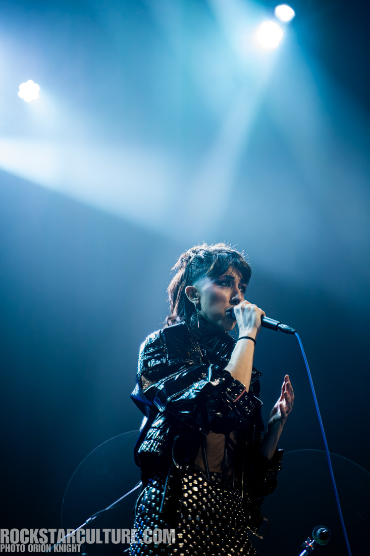 A female singer performing on stage, wearing a shiny black outfit with studs. She is holding a microphone and appears to be passionately engaged with the audience under bright stage lights.