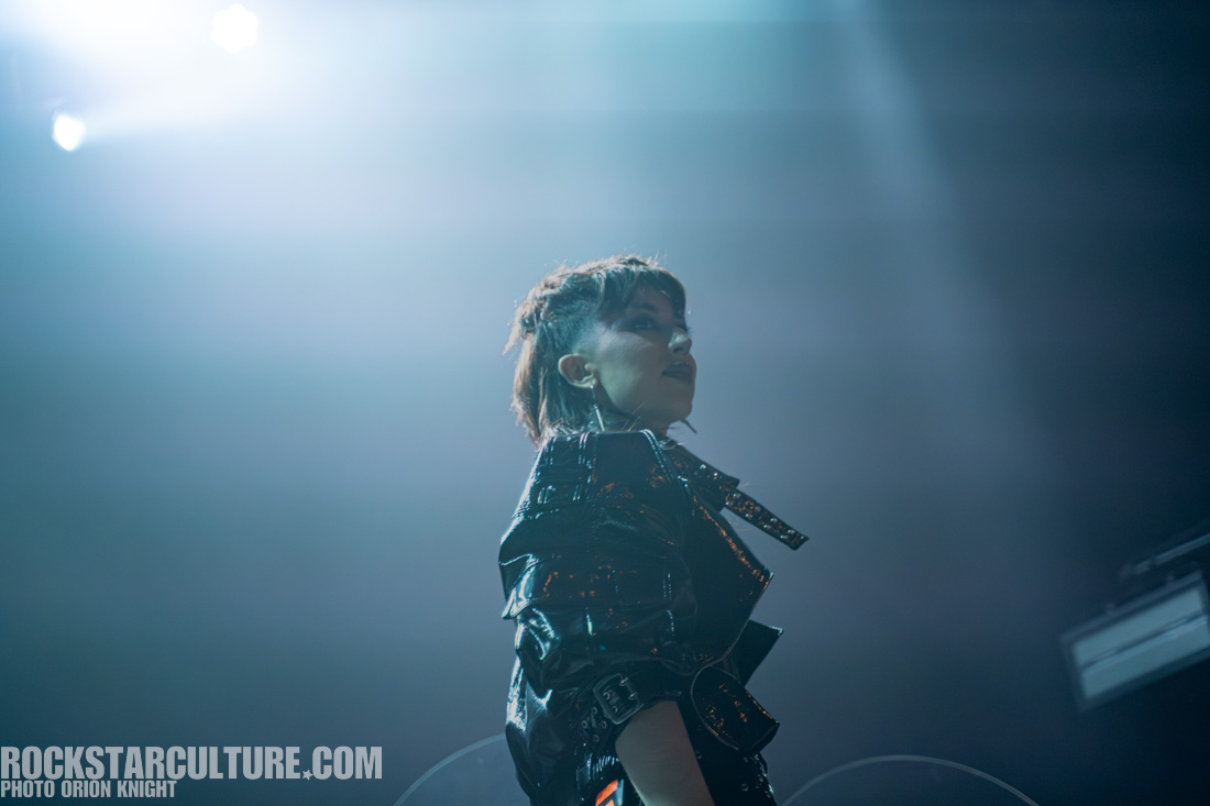 A performer in a shiny black jacket stands in profile under dramatic stage lighting, creating a moody atmosphere.