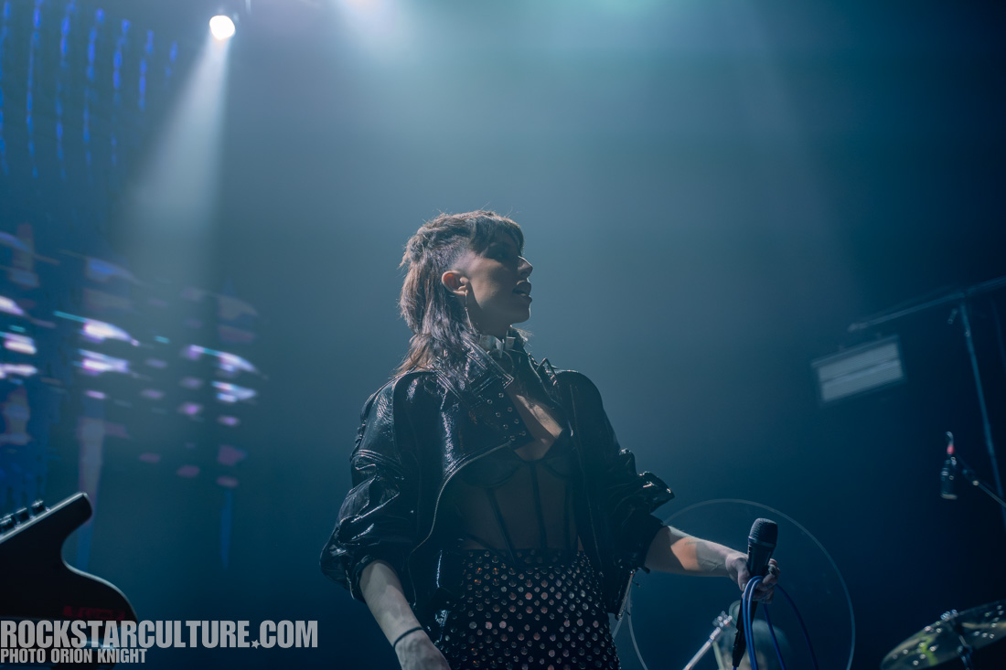 A performer on stage wearing a leather jacket and dotted pants, holding a microphone with a dramatic expression under stage lighting.