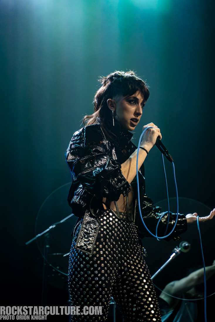 A singer performing on stage, wearing a shiny black outfit with studded pants and holding a microphone. The background features dramatic lighting.