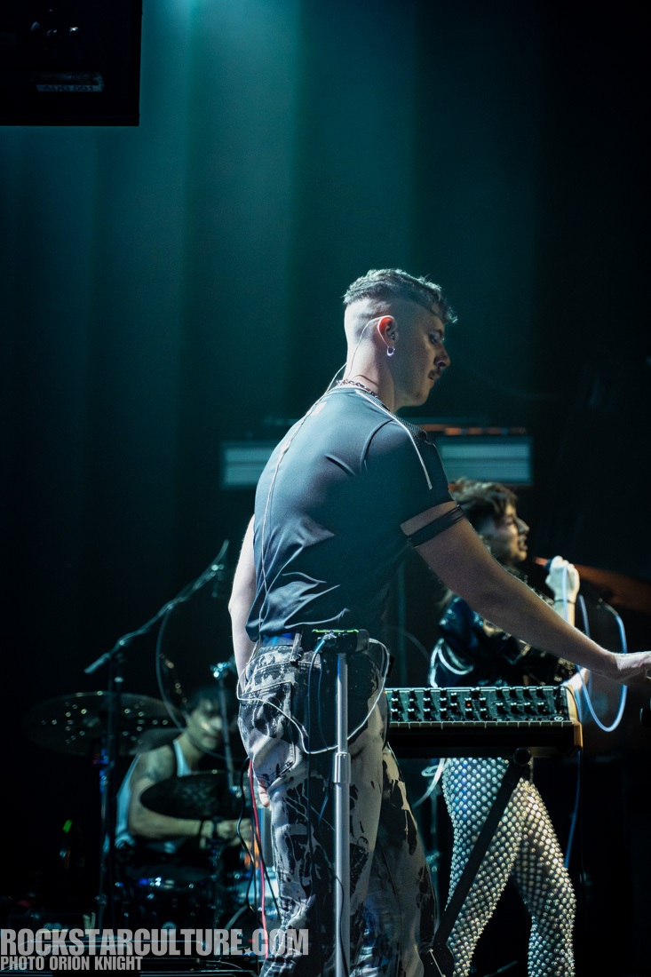 A male musician is performing on stage with a synthesizer, wearing a black shirt and patterned pants, while another band member is in the background playing drums.
