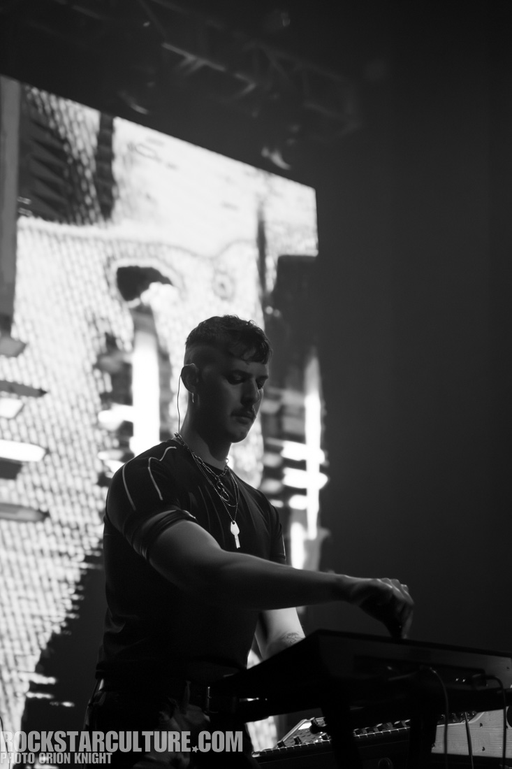 A black and white image of a male musician performing on stage, focused on a synthesizer or keyboard, with a digital backdrop in the background.