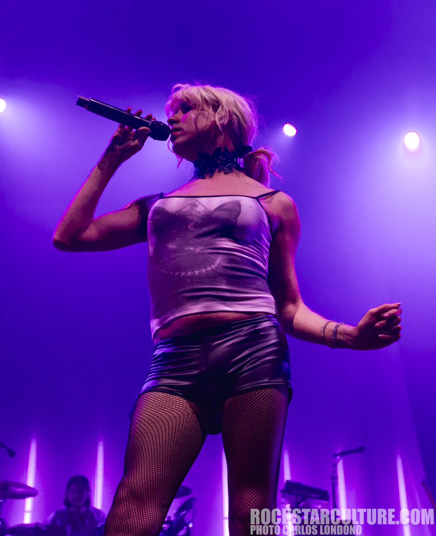 A female performer singing into a microphone on stage, wearing a fitted top and shorts, with fishnet stockings, under purple stage lights.