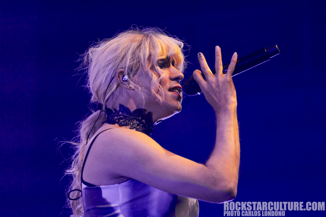 A performer singing into a microphone, wearing a sleeveless outfit and a black lace choker, with blonde hair styled in loose waves against a blue-lit background.
