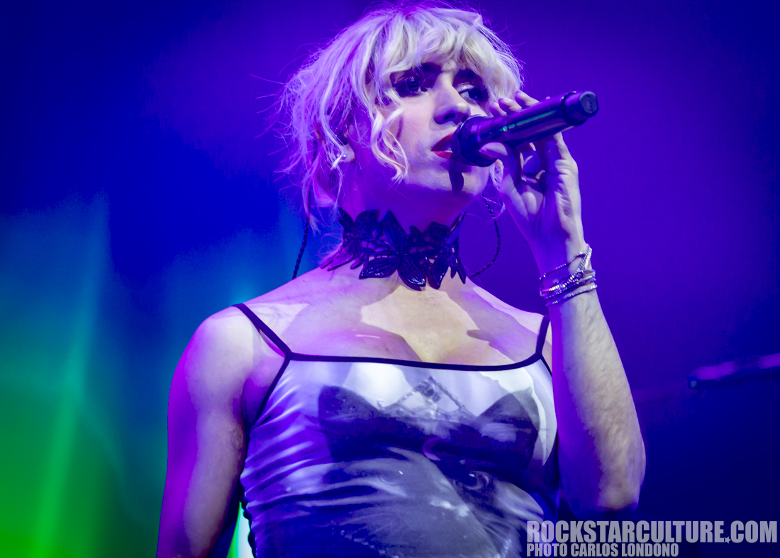 A performer singing into a microphone on stage, wearing a sleeveless dress with a graphic design and a black choker, against a vibrant purple and green background.