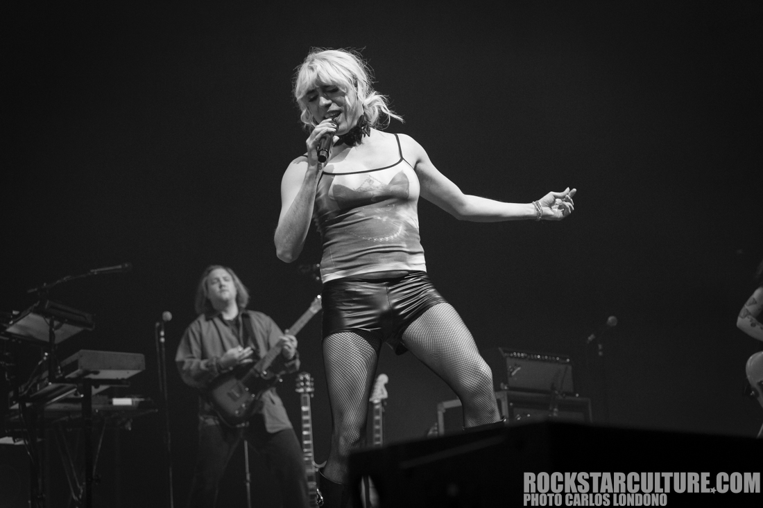 A performer on stage singing passionately while wearing a fitted top and shorts, surrounded by musicians in a concert setting.
