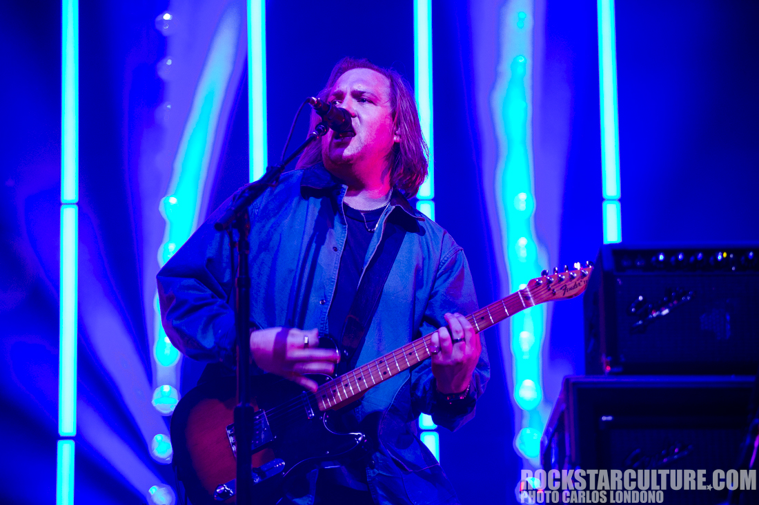 A musician performing on stage with an electric guitar, wearing a blue jacket, illuminated by colorful lights in the background.