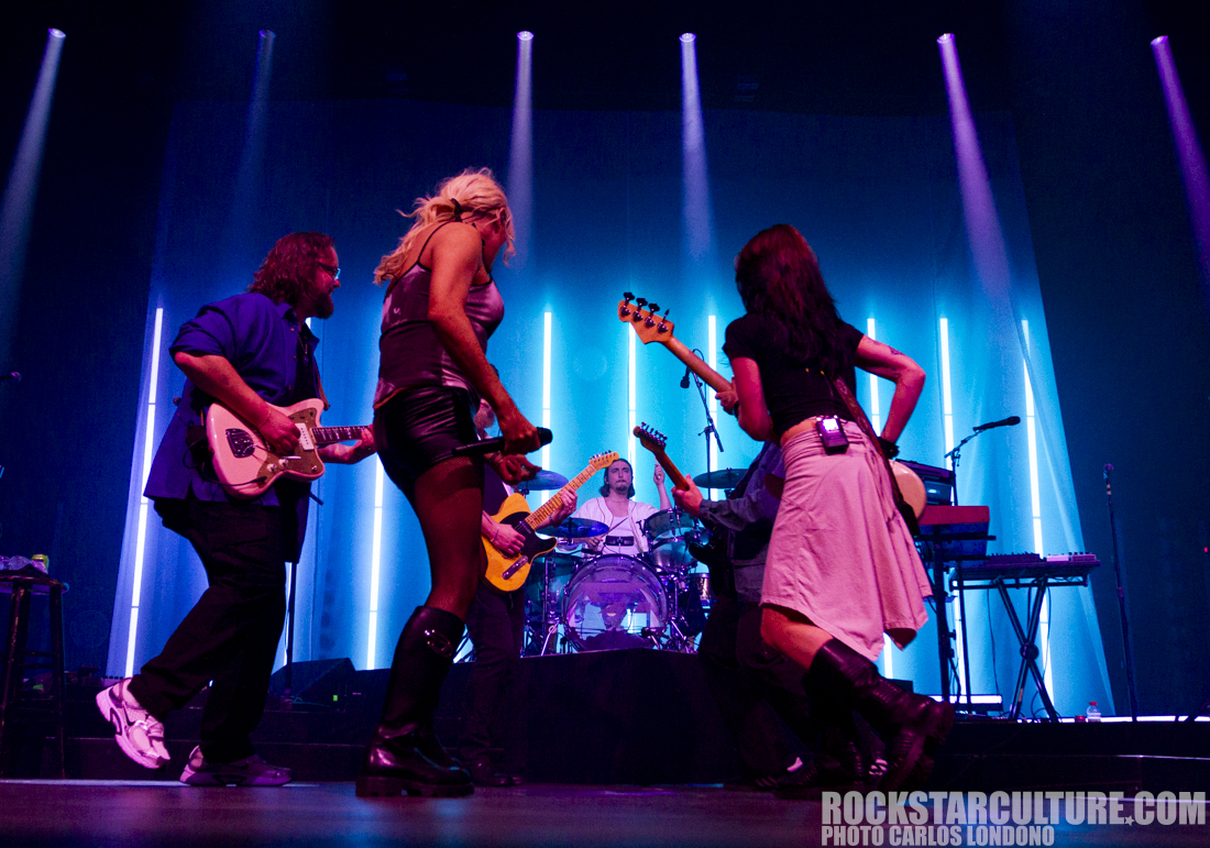 A live music performance featuring multiple musicians playing guitars on stage, with colorful lighting in the background.