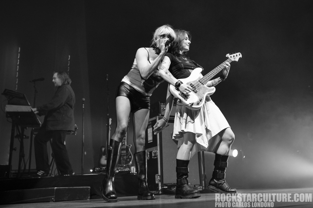 A female vocalist performs energetically on stage, interacting with a female bassist who is playing a white guitar. A male musician can be seen in the background, engaged with a keyboard. The image is captured in black and white, emphasizing the dynamic performance and stage presence.