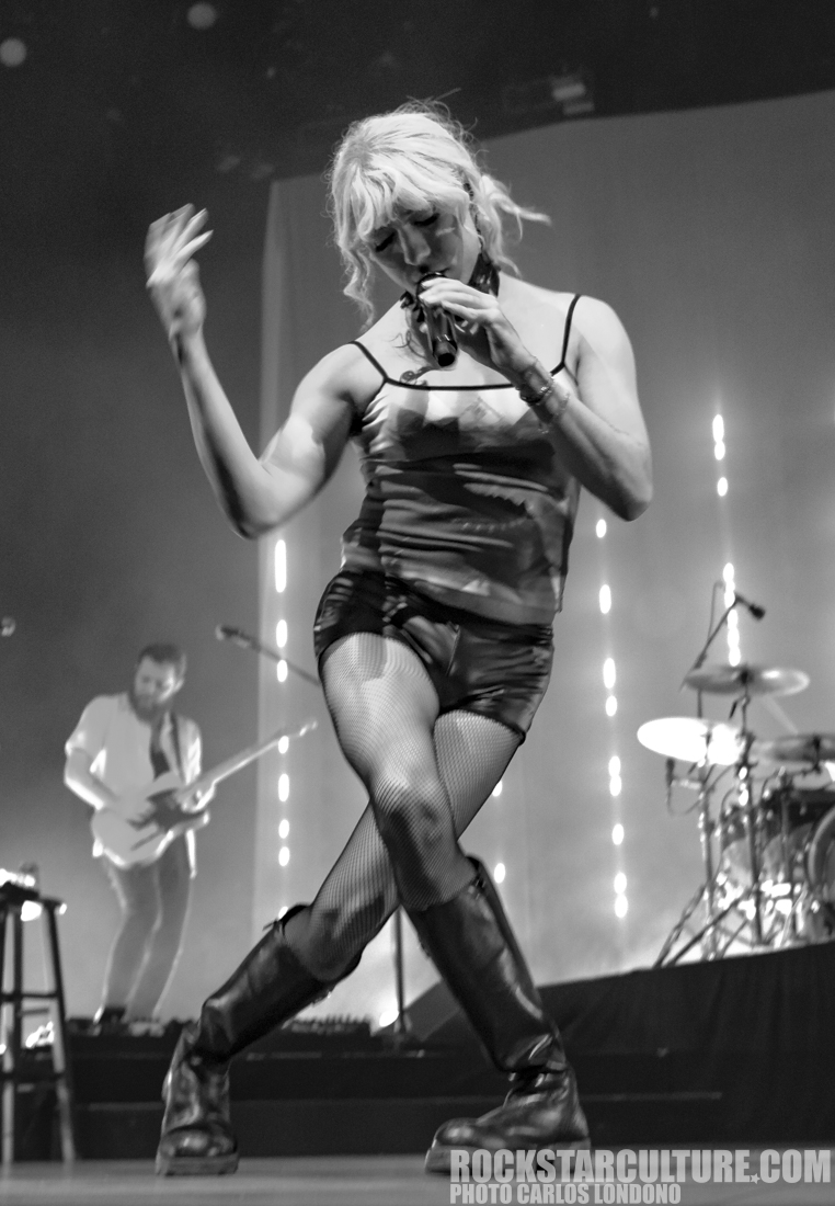 A female singer performing on stage, wearing a black outfit and fishnet stockings, with one hand holding a microphone. The background features a male guitarist and illuminated lines.