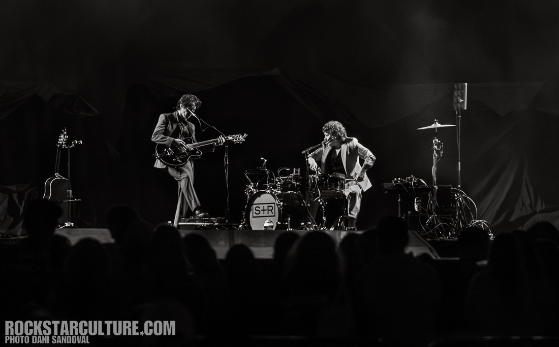 Black and white photo of two musicians performing on stage, one playing electric guitar and the other on drums, with an audience in the foreground.