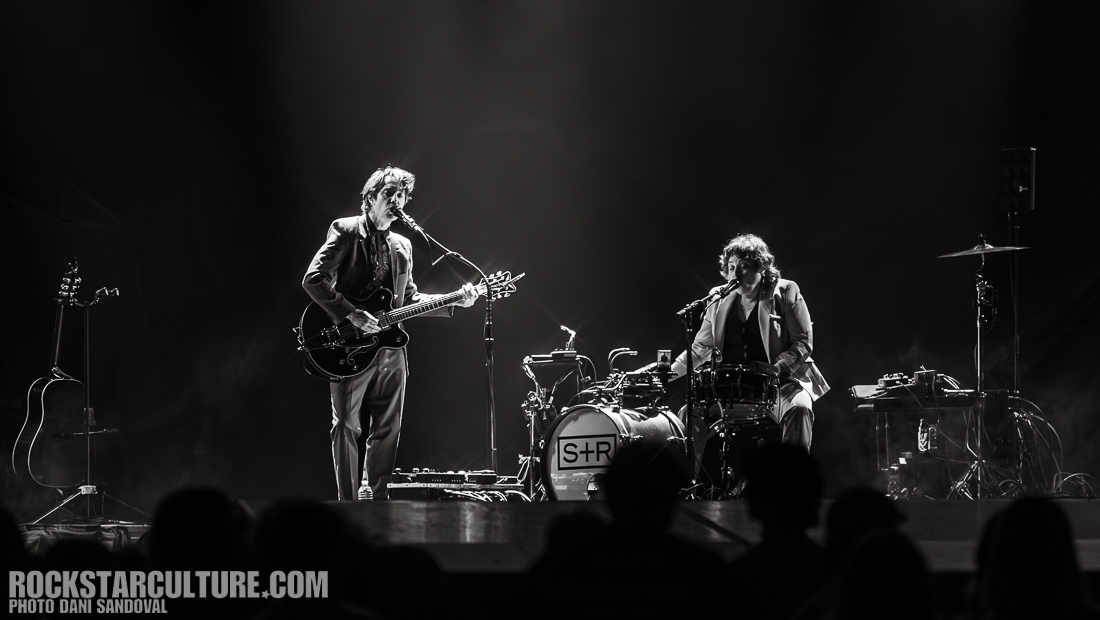 Two musicians performing on stage, one playing electric guitar and the other sitting behind a drum set, both dressed in suits, with a dark background and audience silhouettes visible.