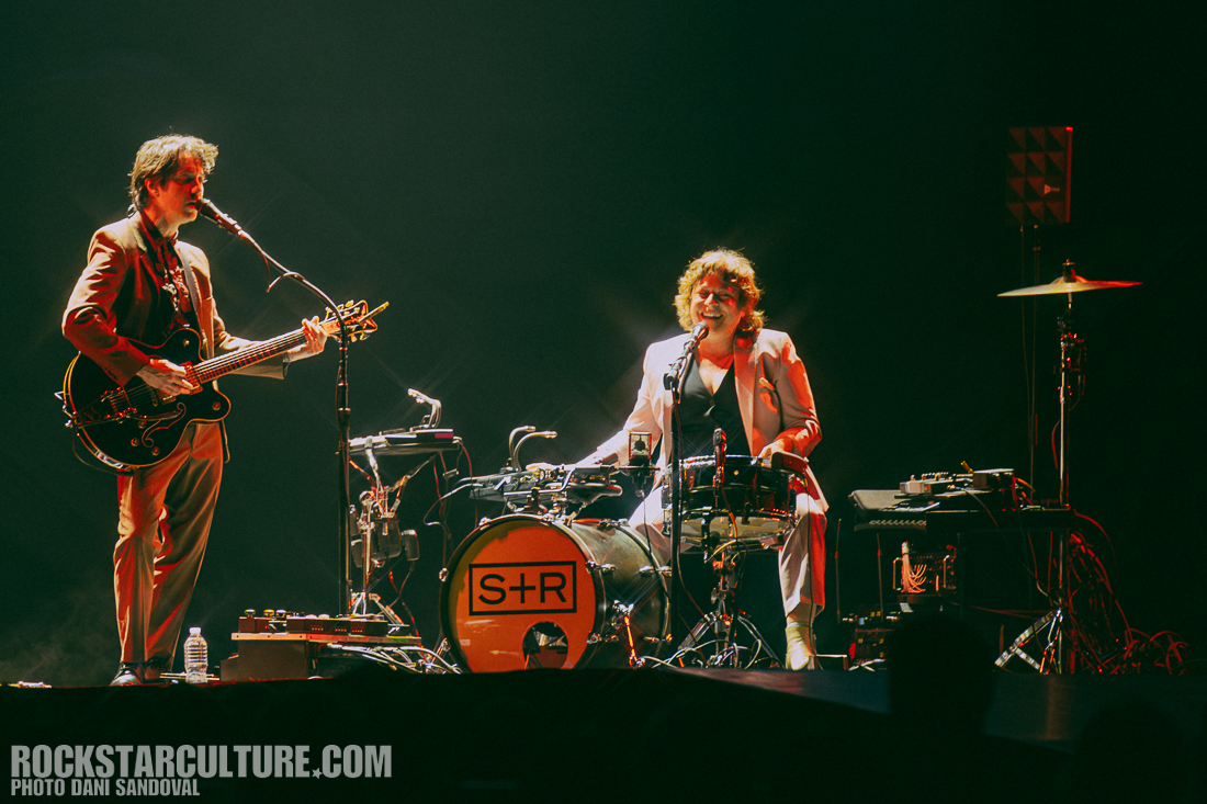 A musician playing an electric guitar and a drummer performing on stage, both smiling and engaging with the audience. The stage is illuminated with focused lighting.