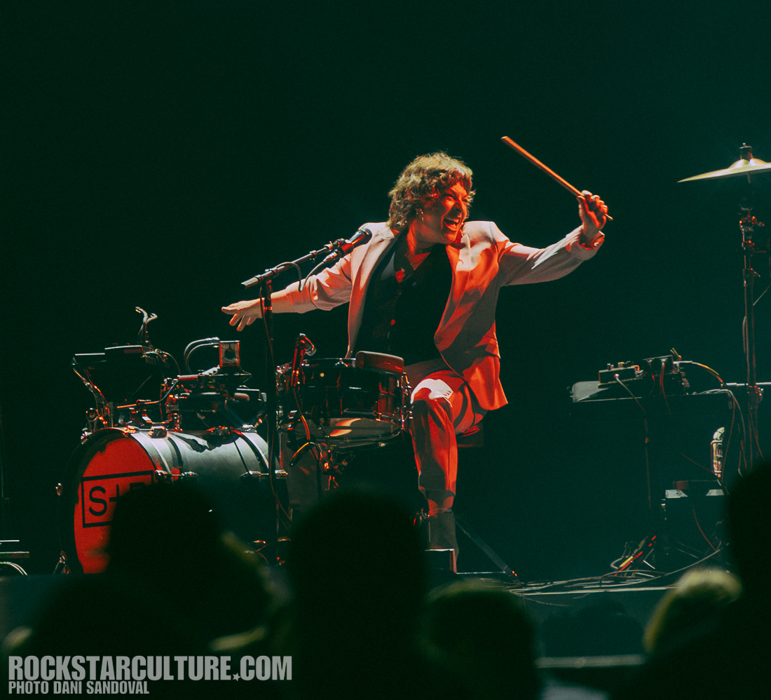 A drummer in a white suit energetically playing the drums on stage, with a microphone and music equipment visible in the background.