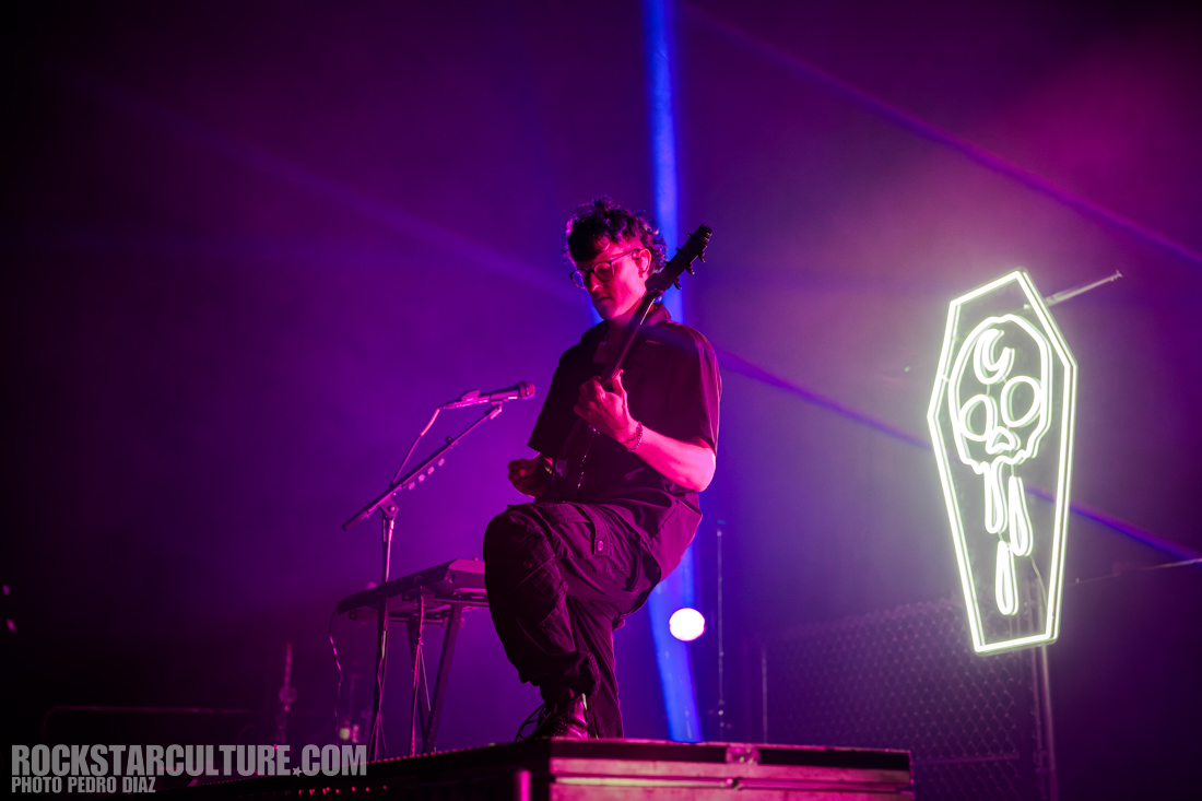 A musician playing an electric guitar on stage, with a keyboard beside him, illuminated by vibrant purple and blue lights. A neon coffin with a cartoon skull design is visible in the background.