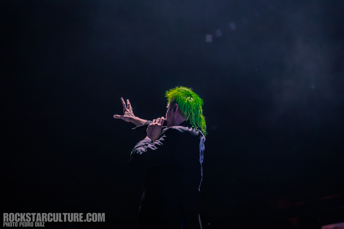 A performer with bright green hair gestures while holding a microphone on stage against a dark background.