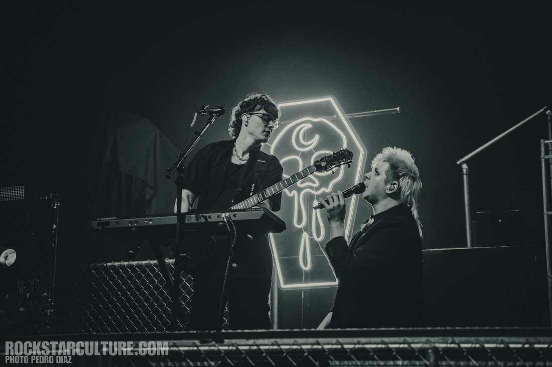 Two musicians performing on stage, one playing a keyboard and the other singing into a microphone, with a neon sign in the background.