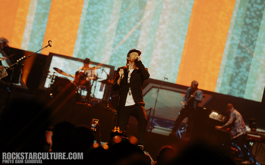 A musician performing on stage with a microphone in hand, surrounded by band members and colorful lighting in the background.