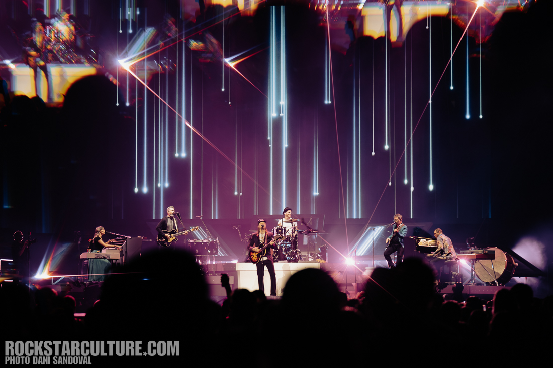 A live band performing on stage with colorful lighting and visual effects, featuring multiple musicians playing various instruments.