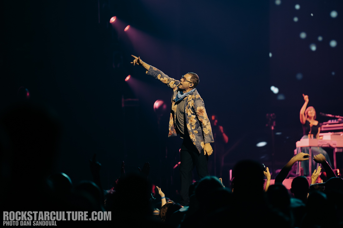 A performer on stage, gesturing with one hand raised while wearing a floral jacket, with a crowd of fans enjoying the show in the foreground.