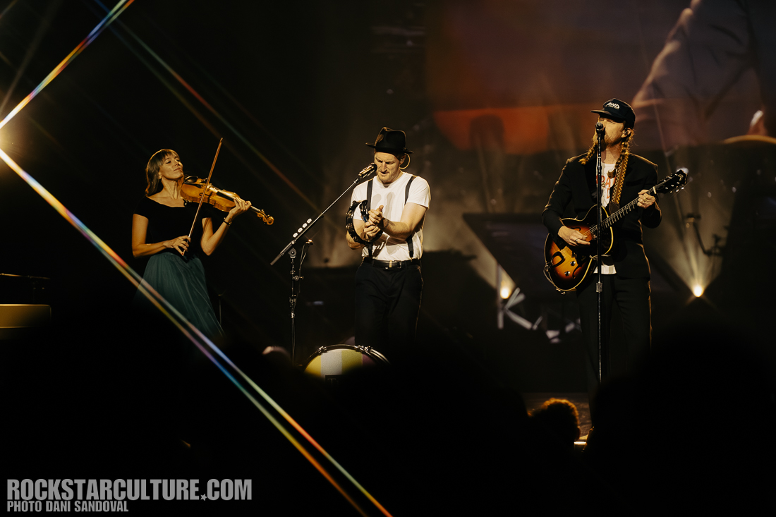 A trio of musicians performing on stage; a female violinist on the left, a male singer playing tambourine in the center, and a male guitarist on the right, with colorful stage lighting in the background.
