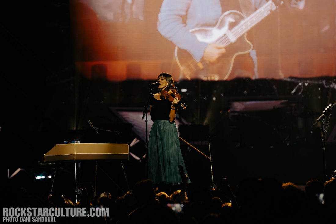 A female musician performing on stage, playing the violin and singing, with a grand piano nearby and an audience in front of her.