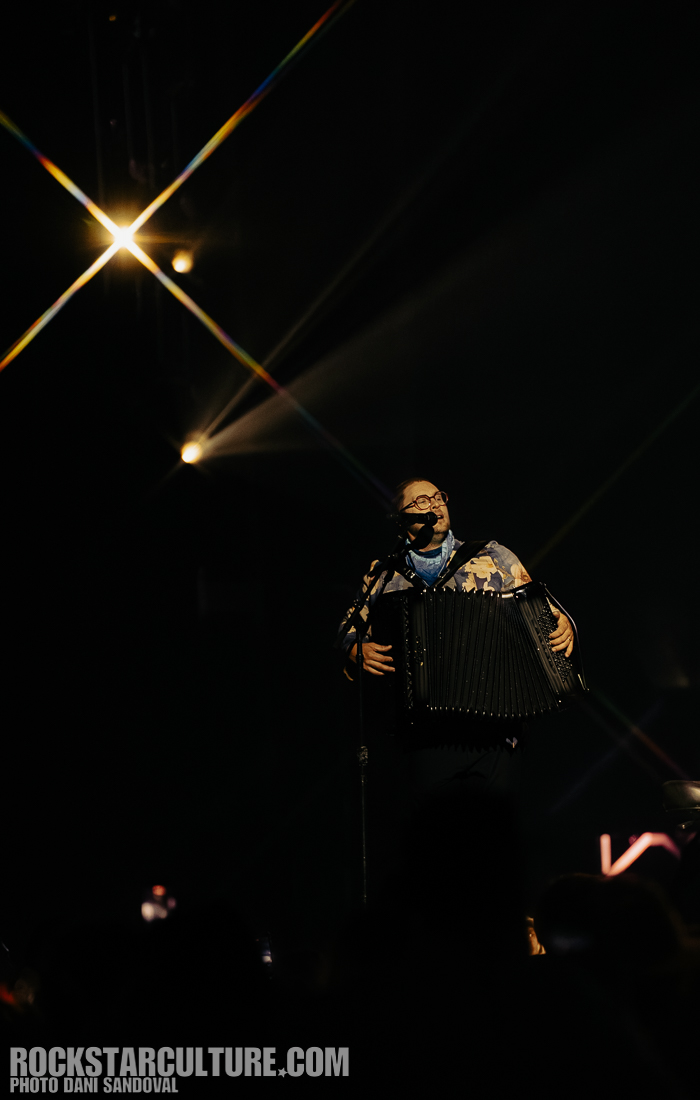 A musician performing on stage with an accordion, wearing glasses and a patterned jacket, illuminated by stage lights.