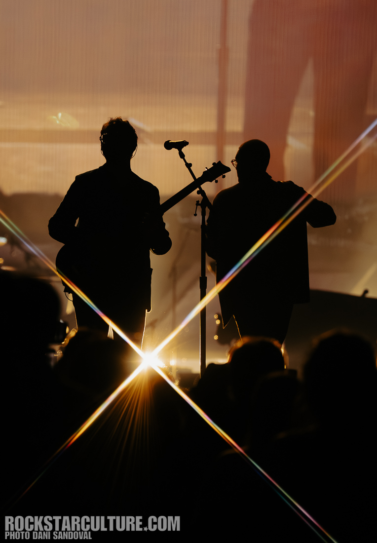 Silhouettes of two musicians on stage, illuminated by bright lights and starburst effects, with an audience visible in the foreground.